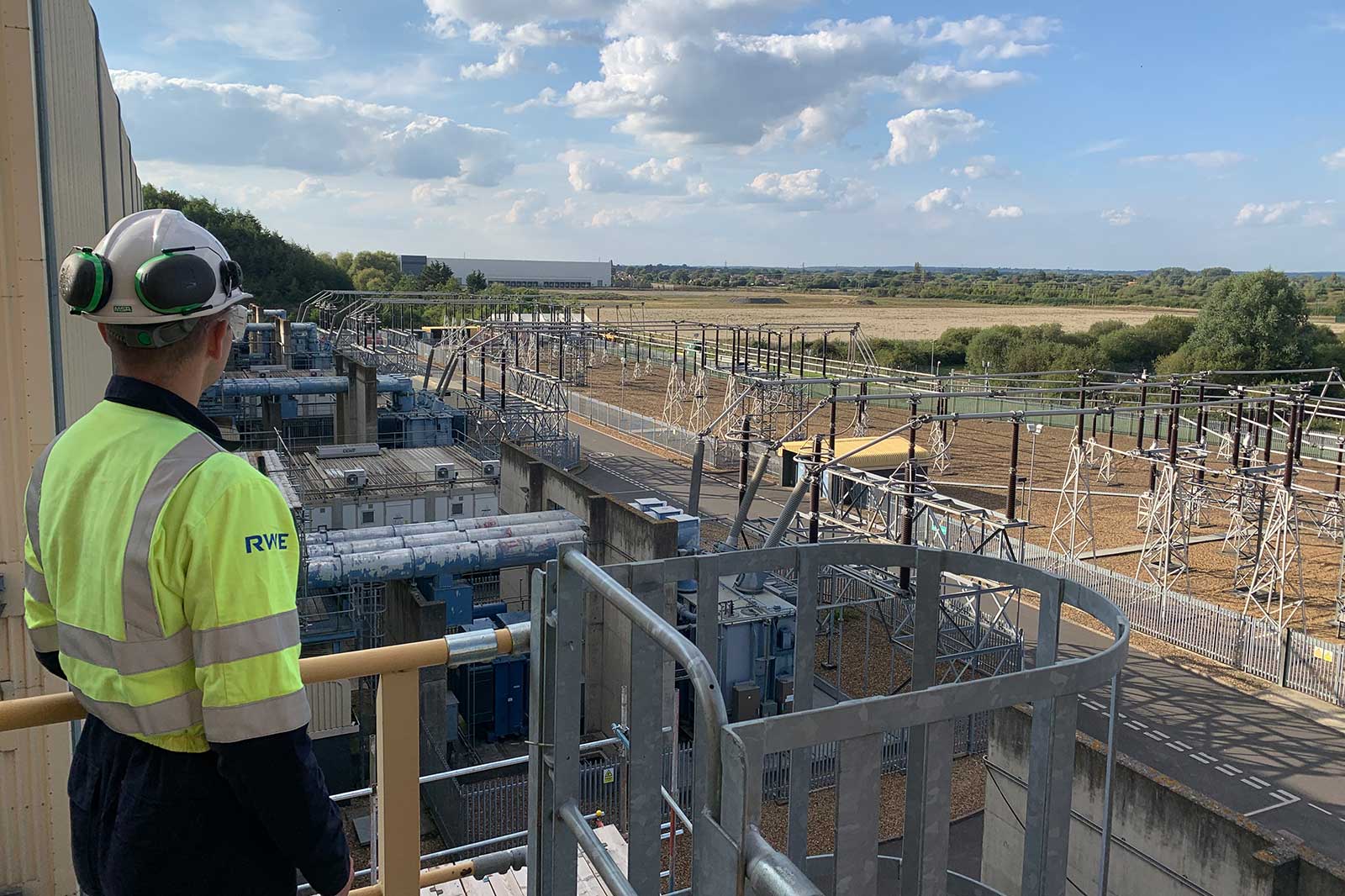 Worker in a high-visibility jacket and helmet overlooks an electrical substation with transformers, pylons and fenced switchgear under a blue sky.