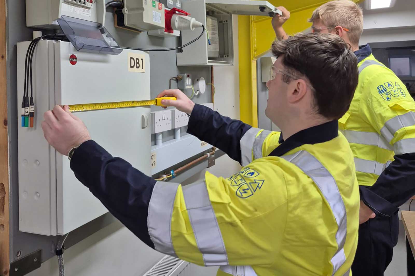 Two workers in high-visibility yellow jackets measure an electrical distribution box labelled DB1 with a tape measure; cables, sockets and reflective stripes are visible.