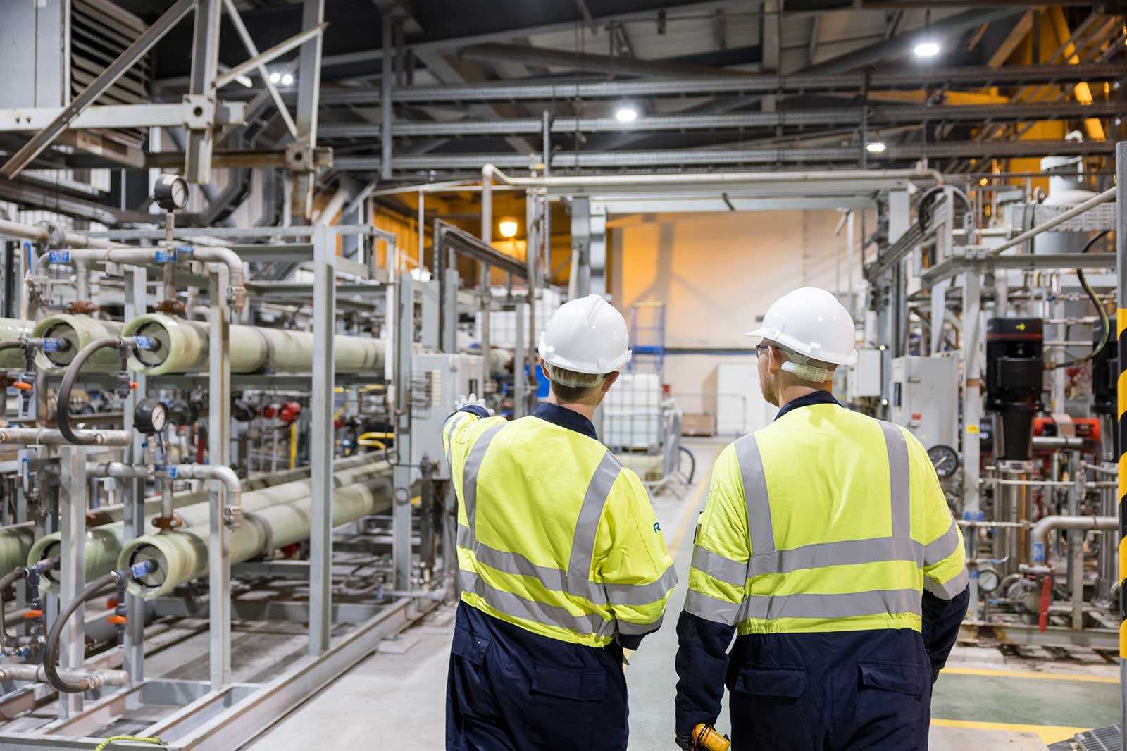 Two workers viewed from behind wearing high-visibility jackets and white hard hats inspect complex pipework in an industrial plant while one points forward.