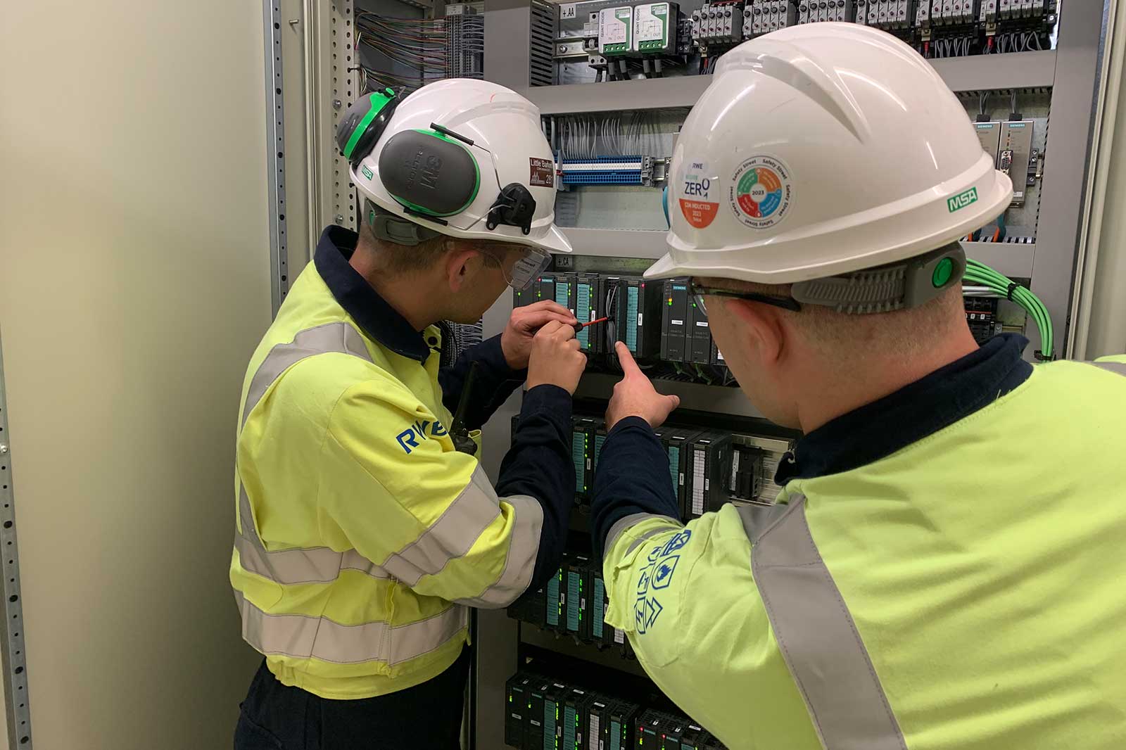 Two workers in hi-vis jackets and hard hats work on an open control panel with PLC modules; one points while the other uses a tool.