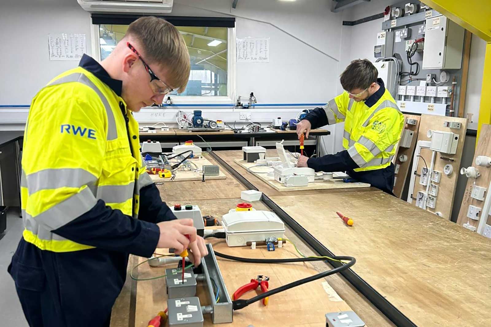 Two technicians in high-visibility RWE jackets work at benches wiring electrical boxes with screwdrivers and tools in a training workshop.