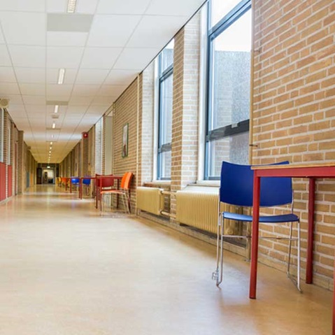 A bright empty school corridor with exposed brick walls, large windows, radiators and colourful chairs and tables along the sides.