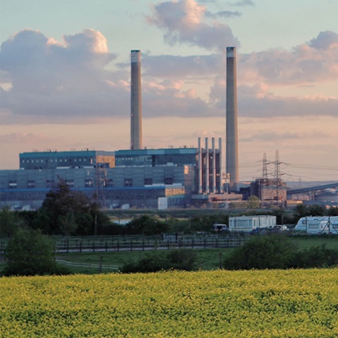A power station with two tall chimneys rises behind yellow flowering fields, caravans and electricity pylons beneath a pastel sunset sky.