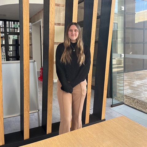 A person stands between vertical wooden slats in a modern space, wearing a black sweater and beige trousers.