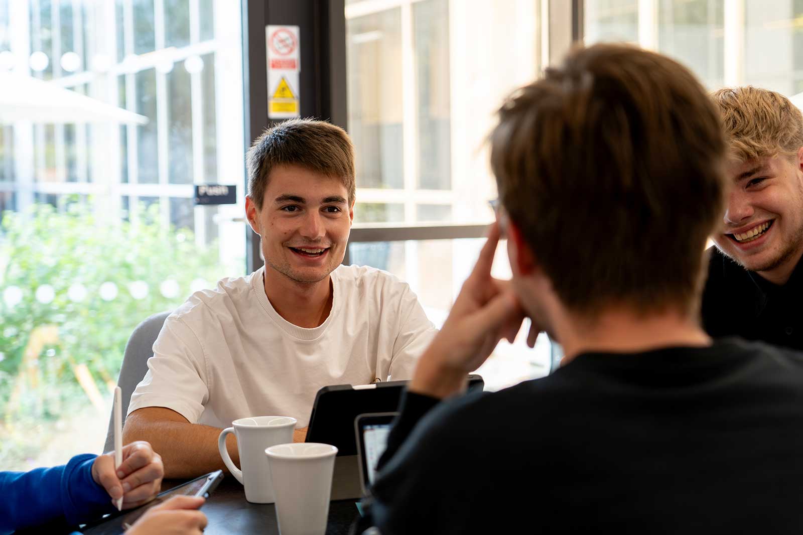 A group of young adults engaged in discussion over coffee at a table with a tablet and cups in a modern café environment.