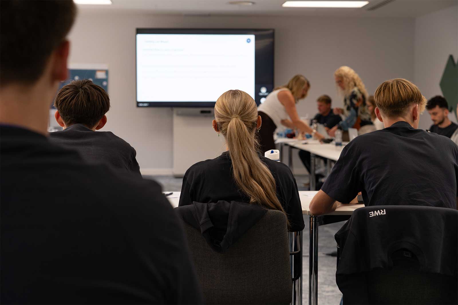 A group of individuals seated in a classroom setting, focused on a presentation displayed on a screen.