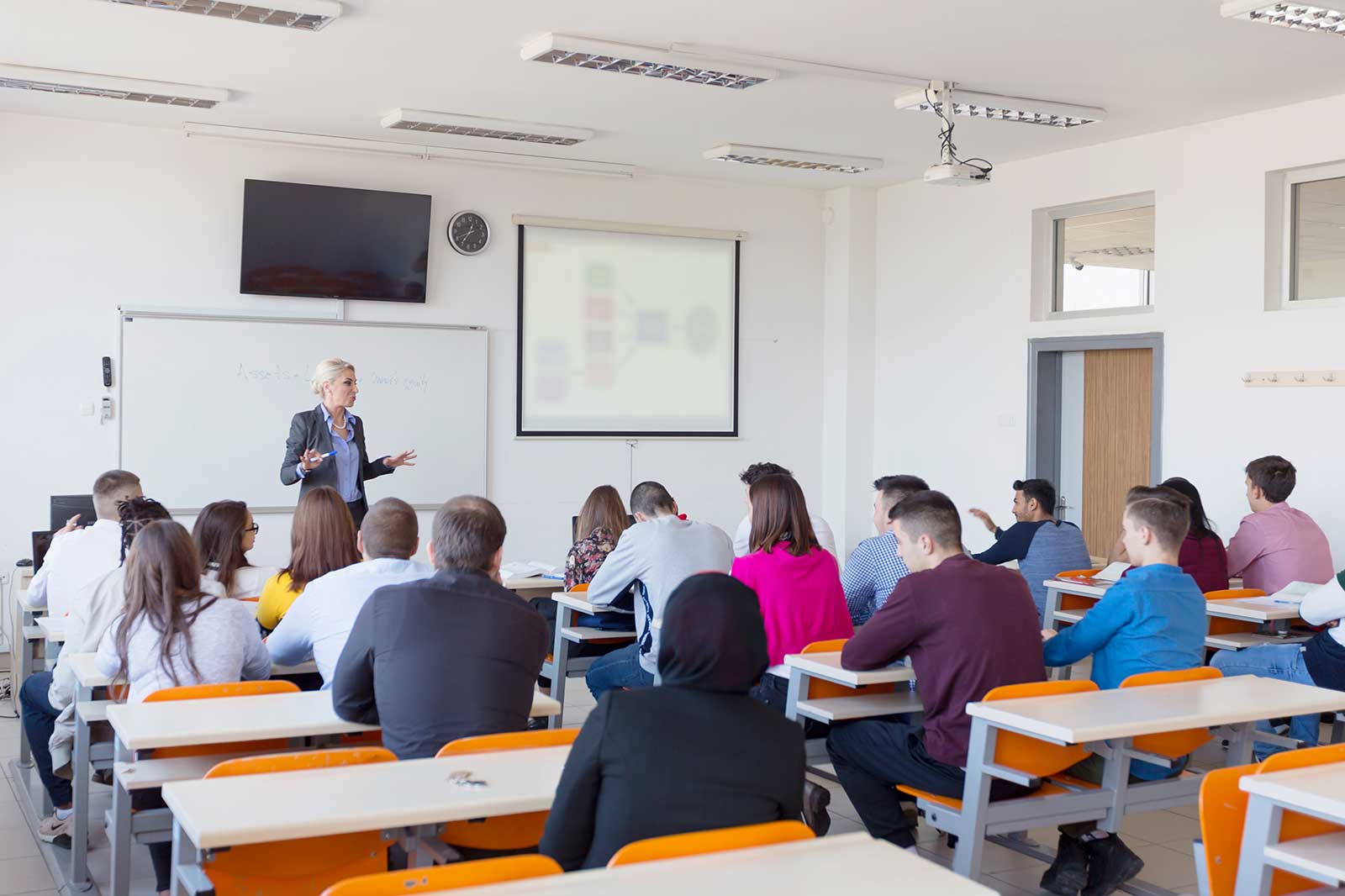 A teacher presents in a bright classroom with students attentively listening at desks with orange accents.
