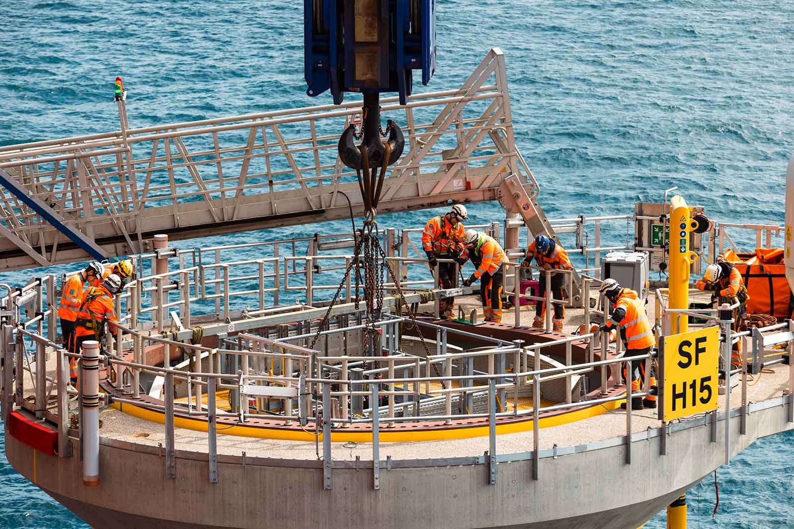 Workers in safety gear are assembling equipment on a circular platform near the sea, with a crane visible overhead.