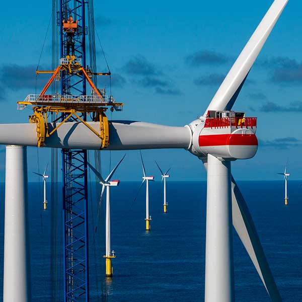A close-up of a wind turbine under maintenance, with a yellow crane on a blue sea and several wind turbines in the background.