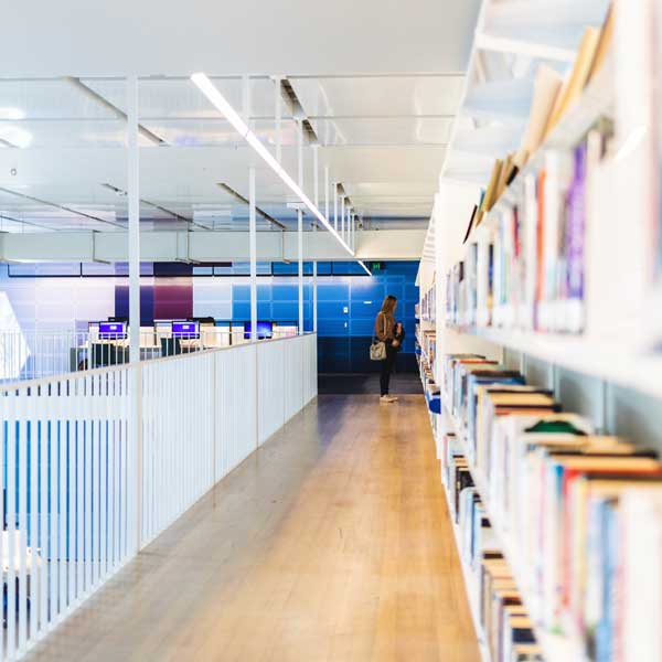 A modern library corridor with bookshelves on the right and a person browsing them, while computers are visible in the background.