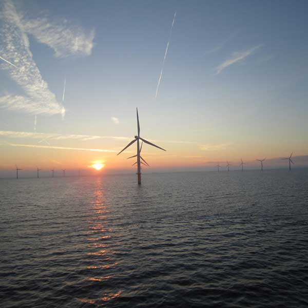 A sunset view over the sea with several wind turbines silhouetted against the horizon and clouds in the sky.