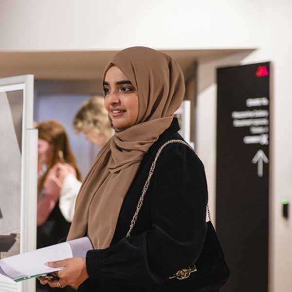 A woman in a brown hijab stands beside an art exhibit, holding a brochure. Other visitors are seen in the background.