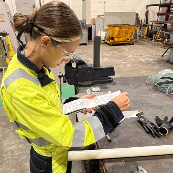 A worker in a yellow safety jacket examines instructions while standing at a workshop table with tools and materials.