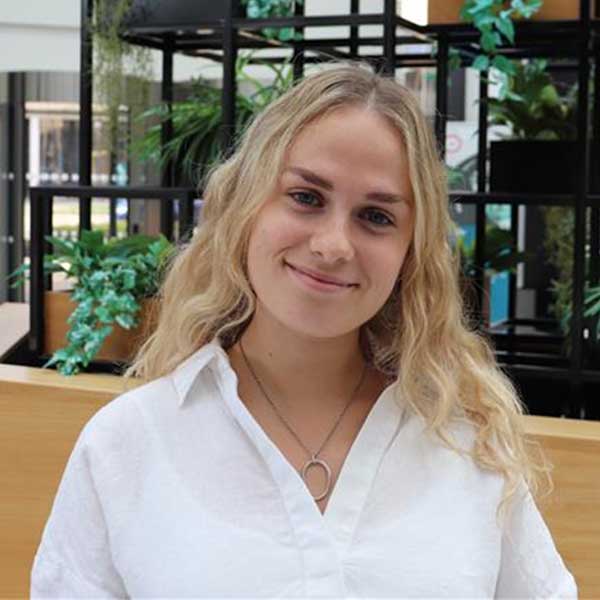 A woman with wavy blonde hair wearing a white shirt stands in front of a green plant backdrop.