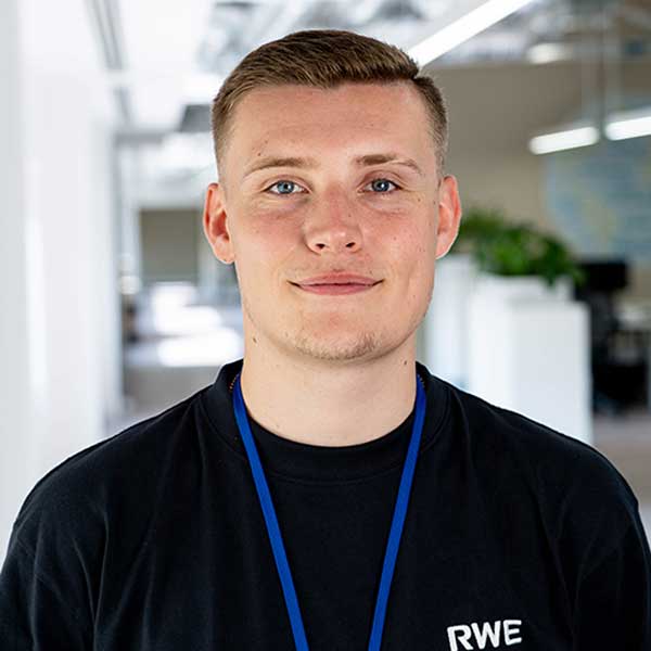 A young man with short brown hair, wearing a black RWE t-shirt and blue lanyard, stands in a modern office setting.