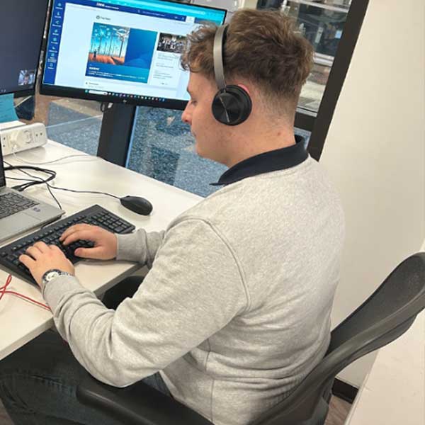 A person sits at a desk with a laptop and monitor, wearing headphones, engaging with a digital interface.