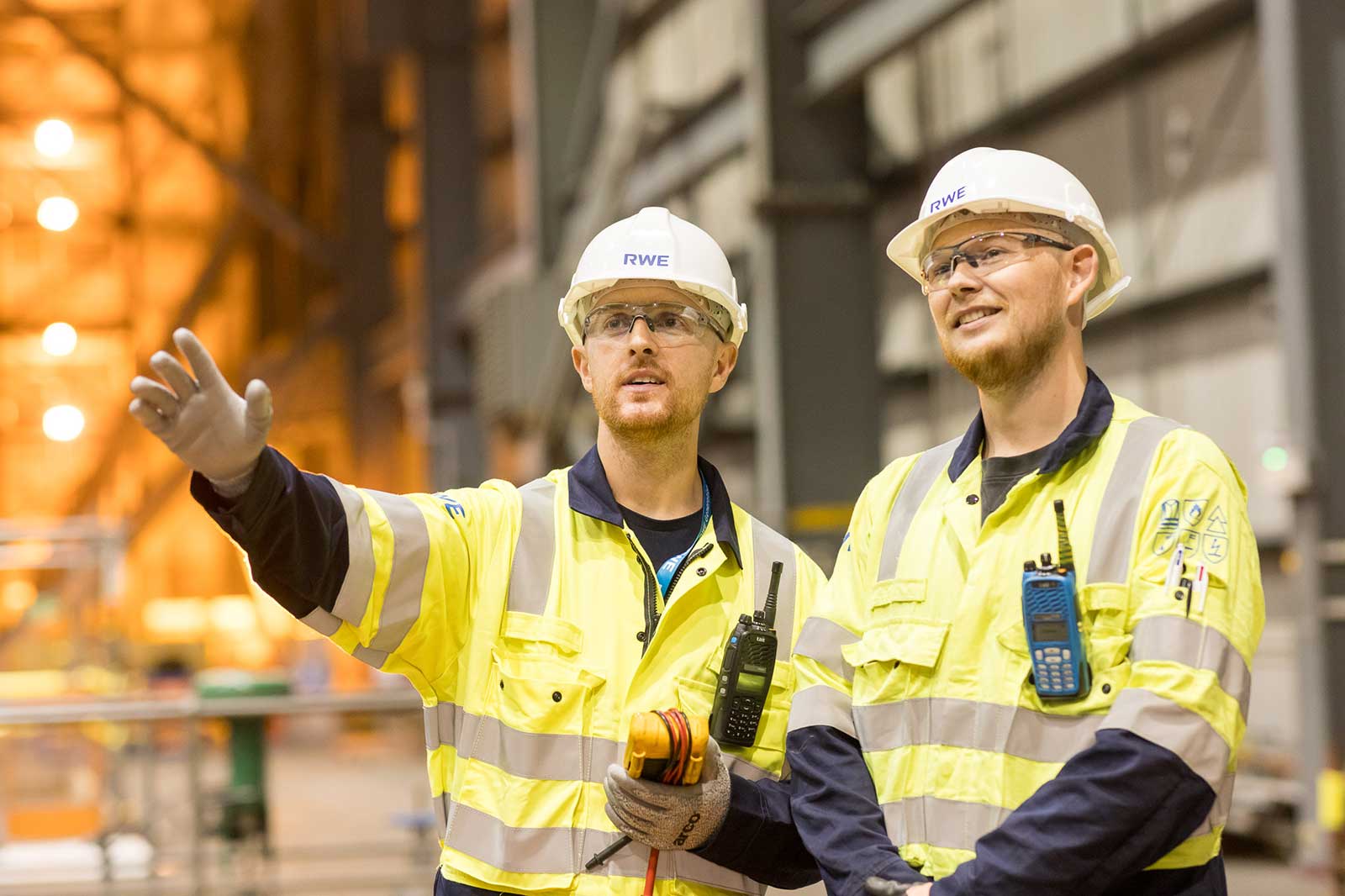 Two workers in high-visibility jackets and helmets, one gesturing and holding tools, in a large industrial space with equipment.