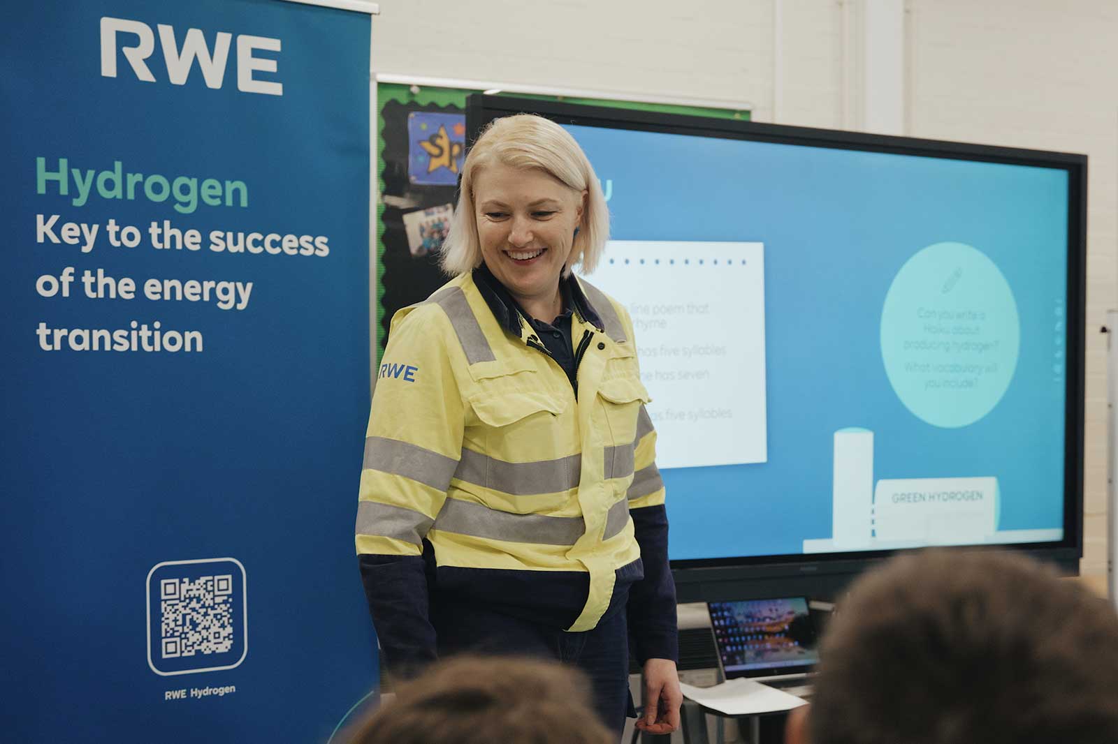 A speaker in a yellow safety jacket stands in front of a presentation about hydrogen's role in energy transition.