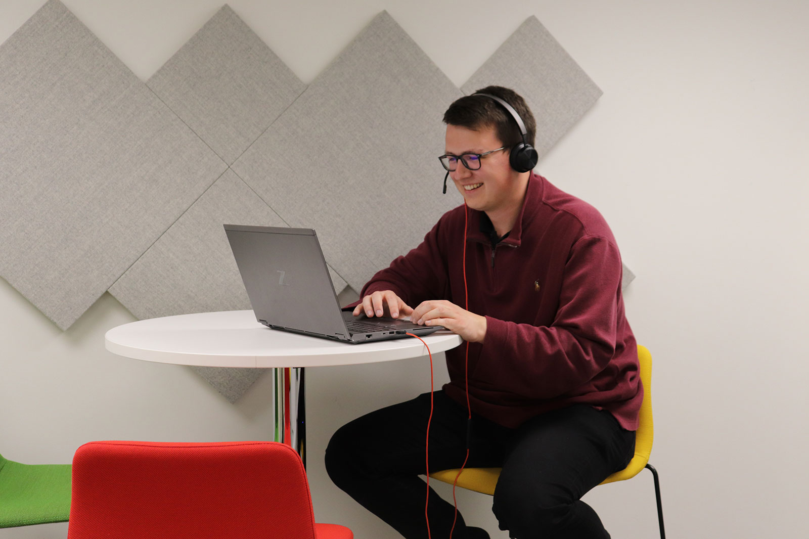 A person in a maroon jumper is working on a laptop at a round white table, wearing headphones in a modern workspace.