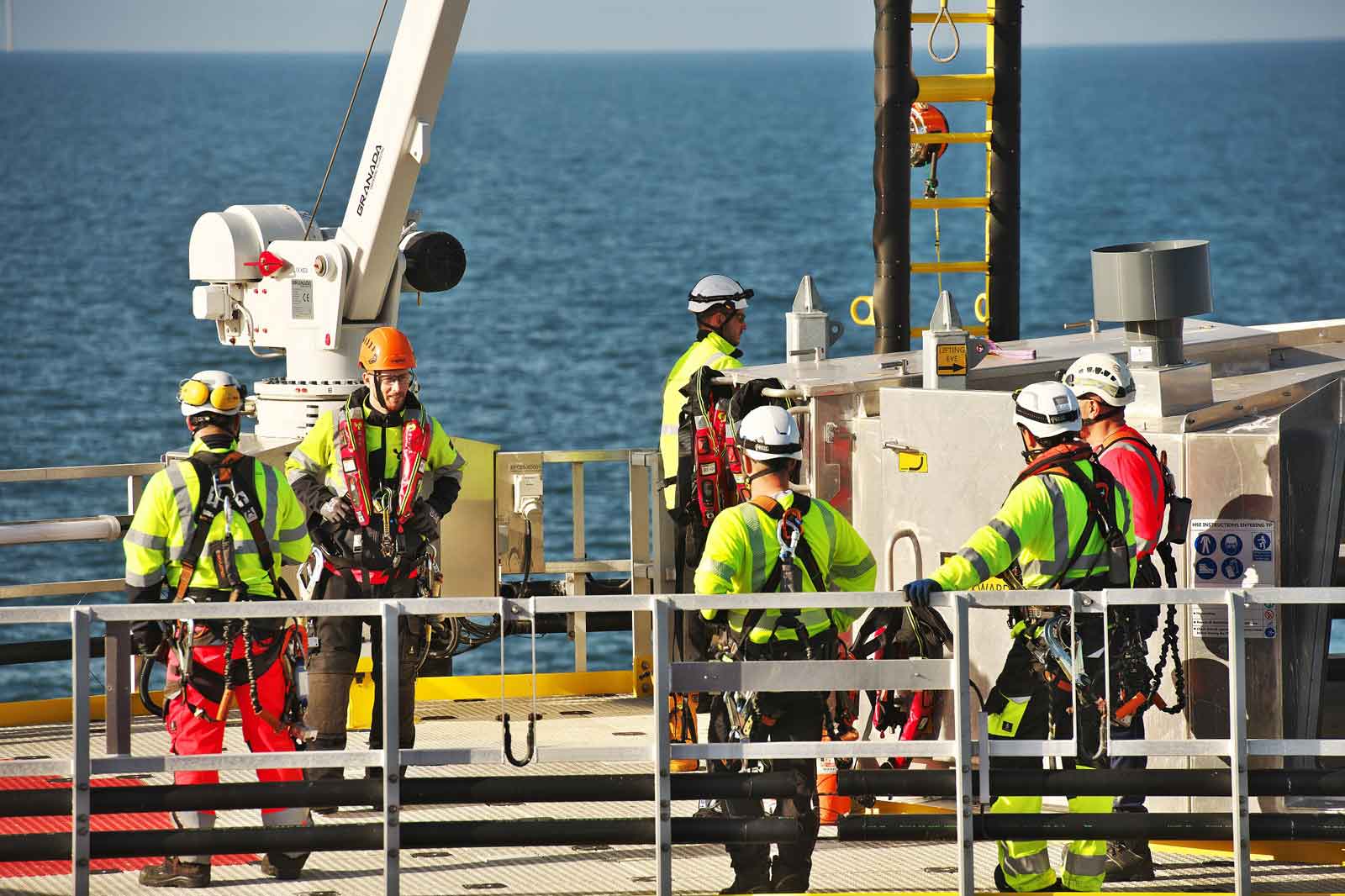 Six workers in safety gear and helmets stand on a platform above the ocean, discussing tasks with equipment in the background.