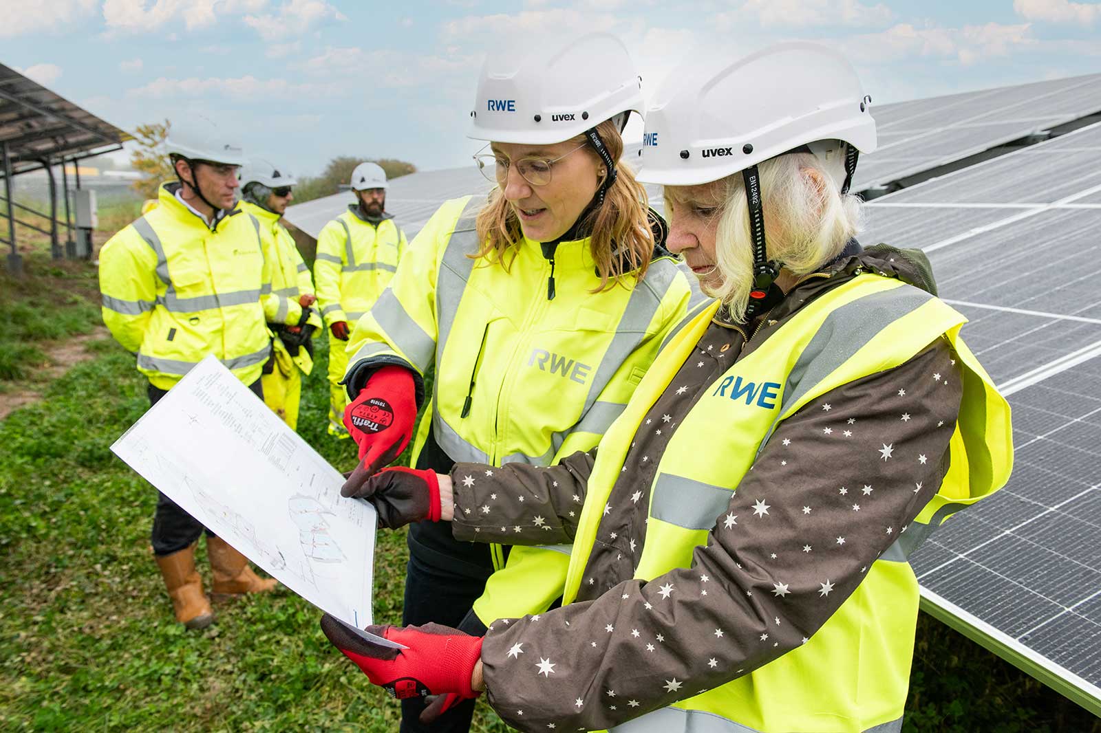 Two workers in neon jackets examine a plan near solar panels, with colleagues in the background.