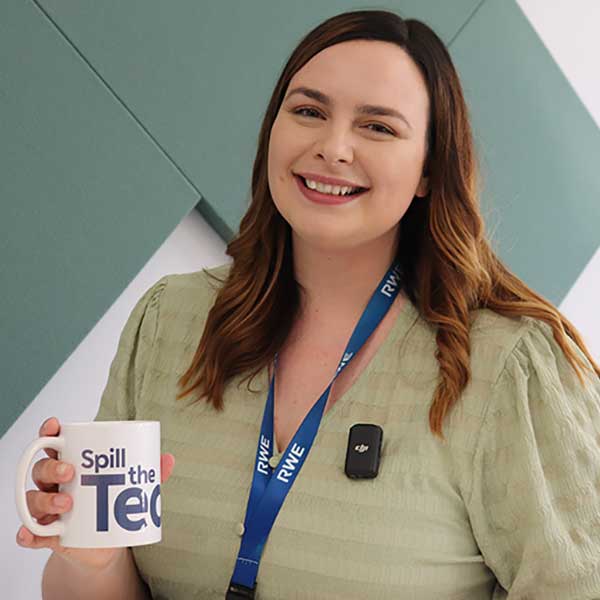 A person in a light green shirt holds a white mug that reads 'Spill the Tea'. A lanyard is around their neck.