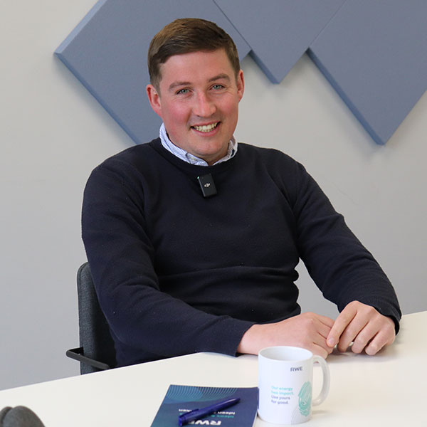 A man sits at a table, wearing a dark sweater with a microphone. A white mug and documents are on the table.