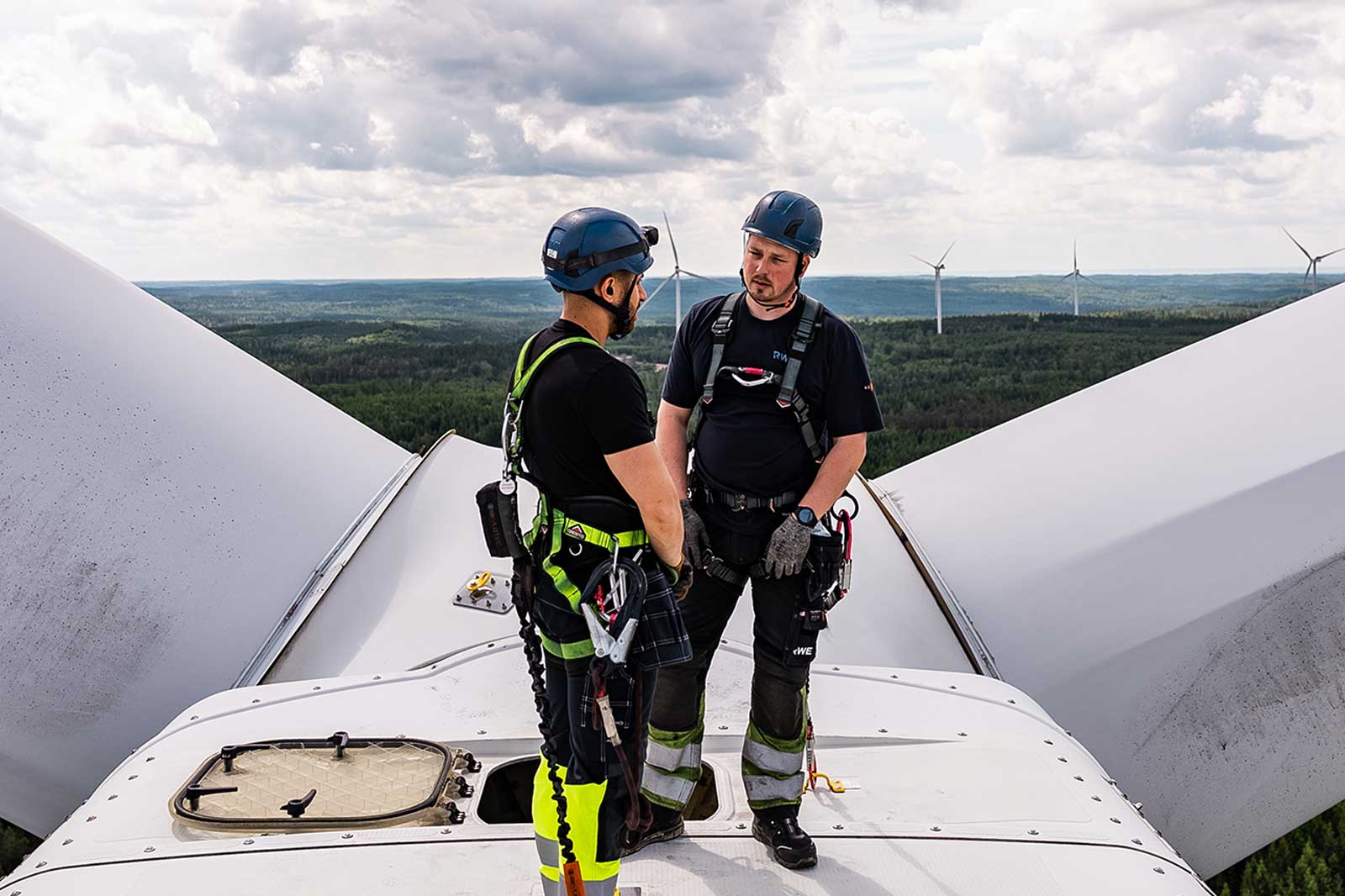 Two technicians wearing helmets and harnesses stand atop a wind turbine, surrounded by a green landscape and wind turbines in the distance.