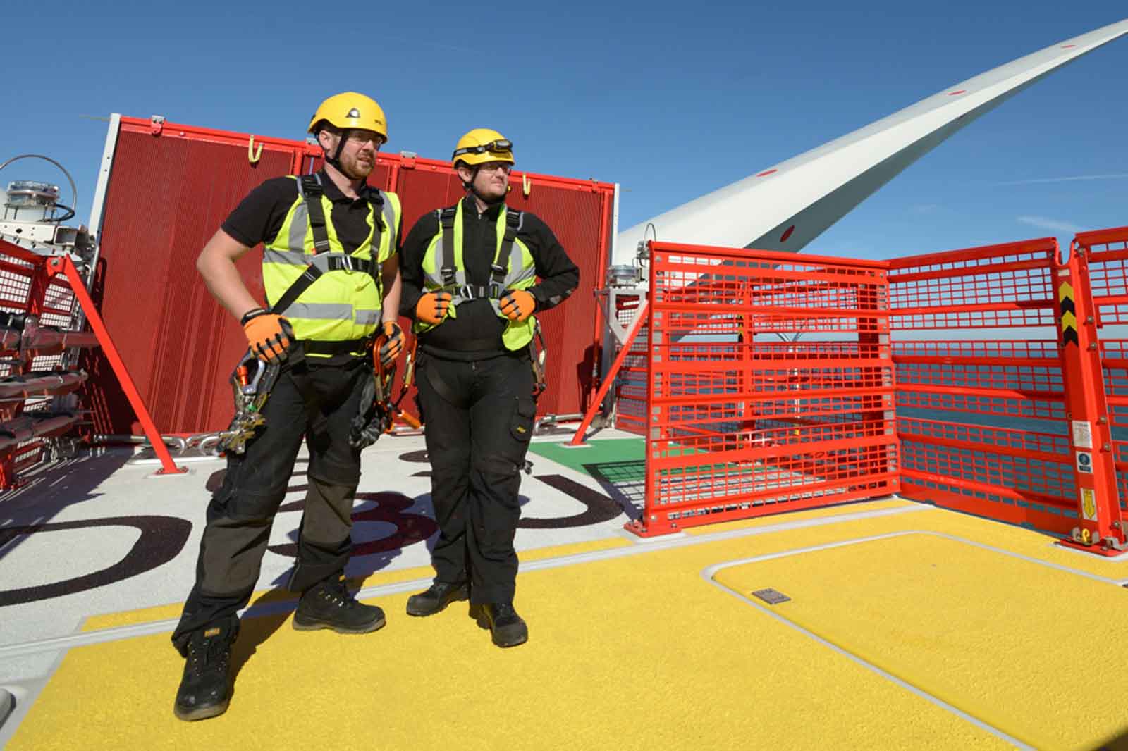 Two workers in safety gear stand on a yellow platform, overlooking wind turbine blades against a clear blue sky.