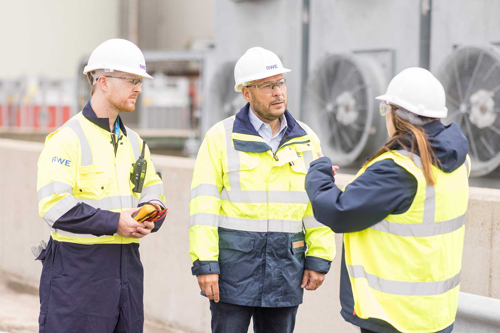 Three individuals in safety gear discussing near industrial equipment with air conditioning units in the background.