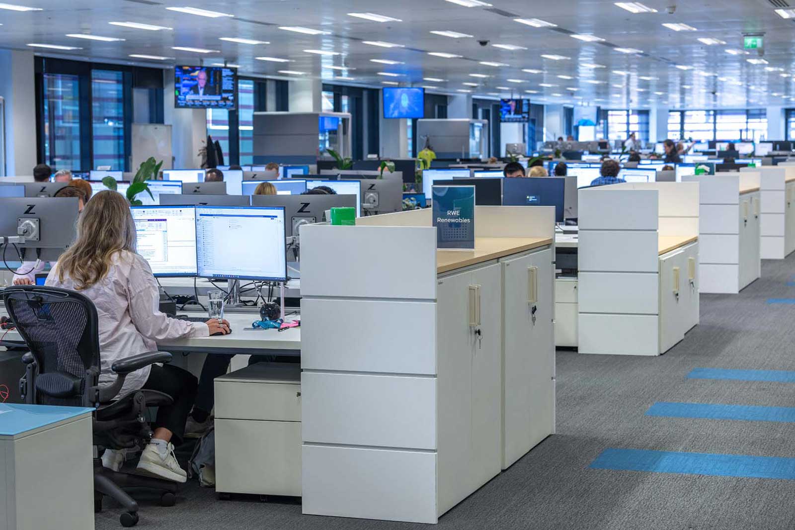 A modern office space featuring multiple workstations with computers, plants, and a presentation screen in the background.