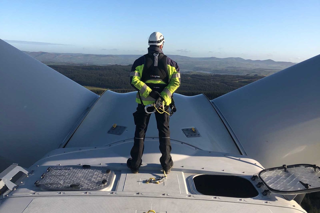 A worker in safety gear stands on top of a wind turbine, overlooking a vast landscape of hills and forests under a clear sky.