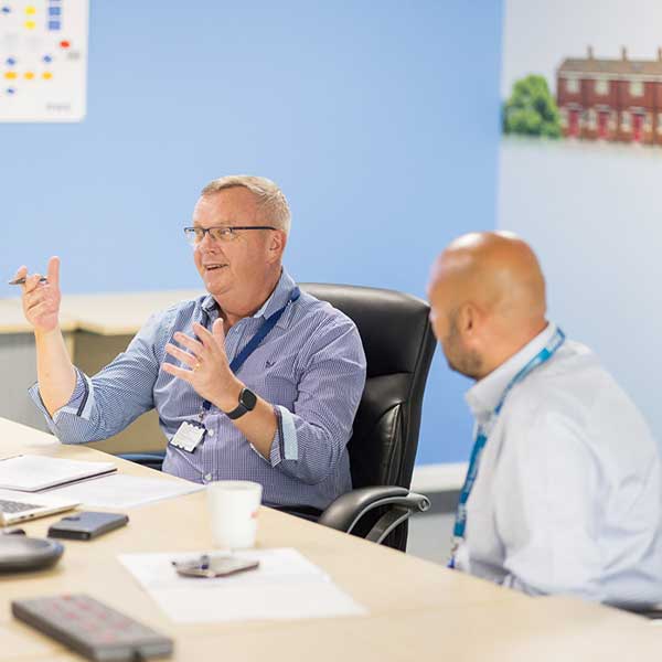 Two colleagues engaged in a discussion at a meeting table with a laptop and documents in a bright office setting.