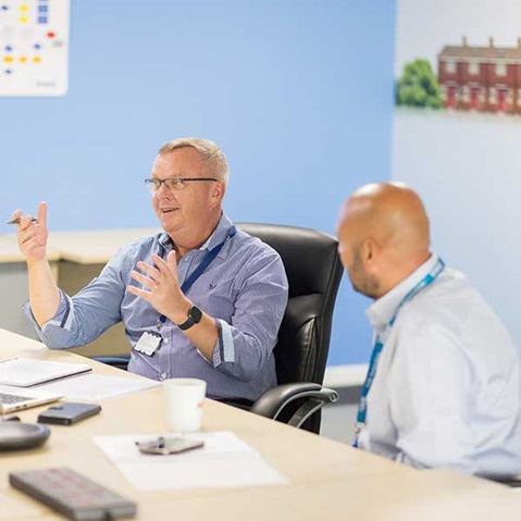 Two colleagues engaged in a discussion at a meeting table with a laptop and documents in a bright office setting.