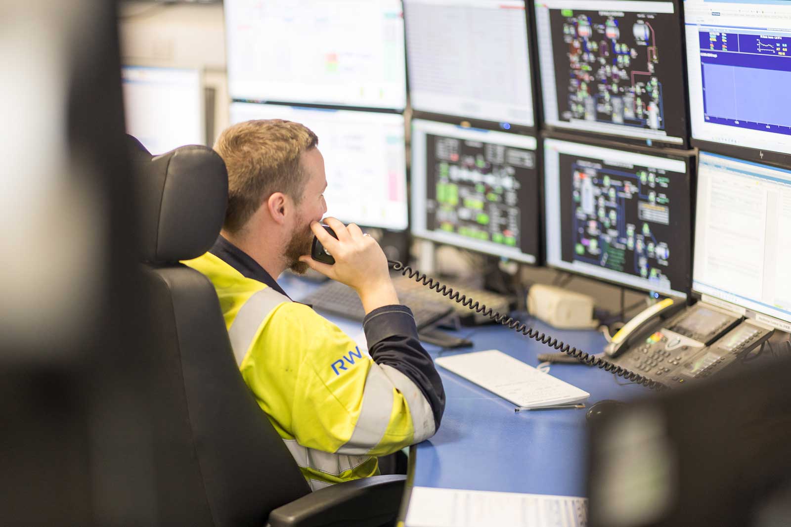 A worker in a safety jacket communicates over the phone while surrounded by multiple computer monitors displaying operational data.