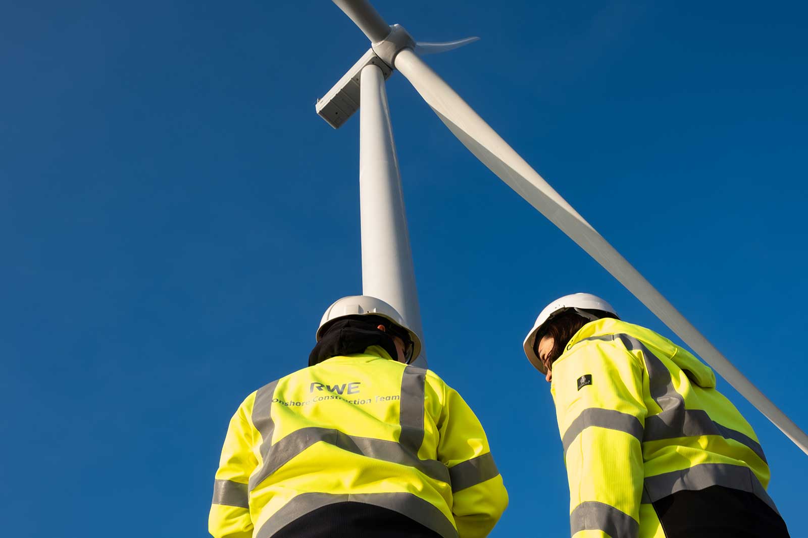 Two workers in reflective jackets and hard hats examine a wind turbine against a clear blue sky.