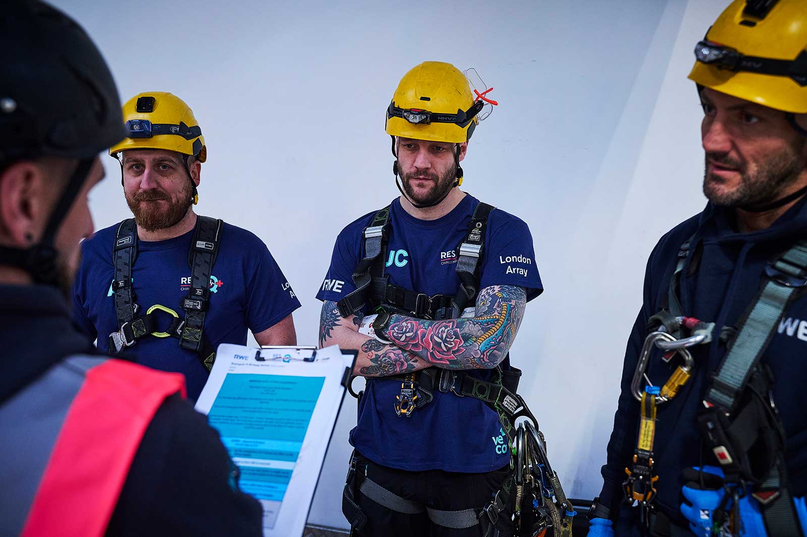 Three climbers in safety gear converse as one holds a clipboard. They wear helmets and harnesses.