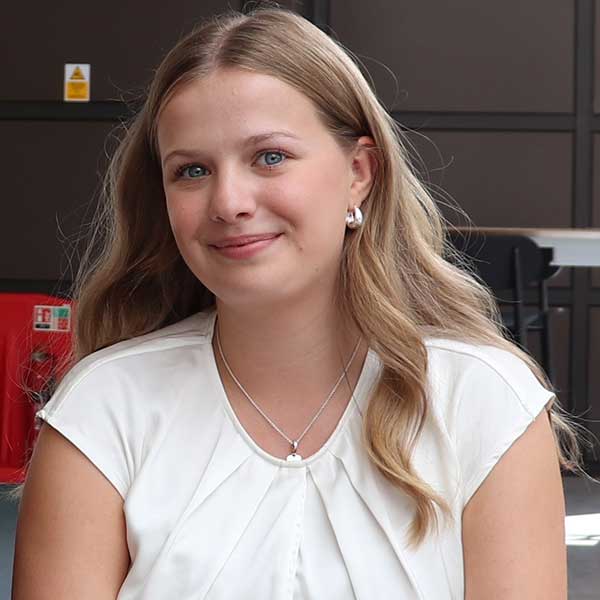 A woman with long wavy hair wearing a white blouse and jewellery, seated in a modern setting.