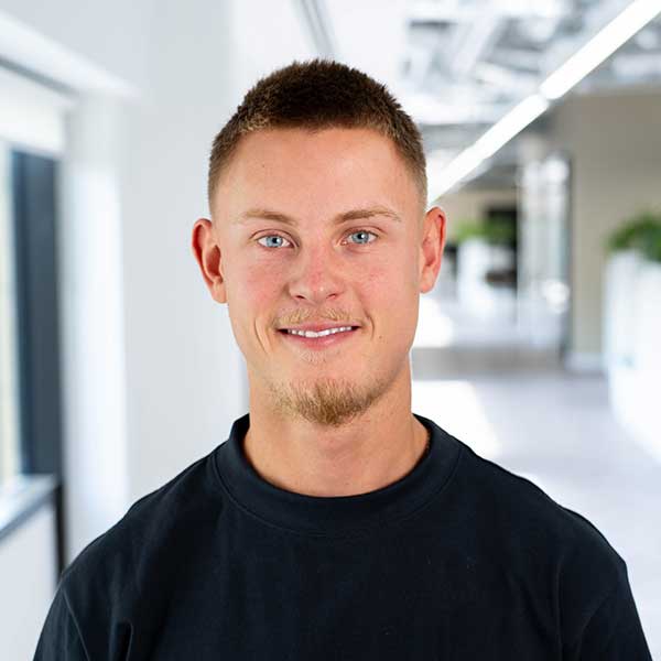A person stands in a modern corridor, wearing a black sweatshirt. The background features bright lighting and plant elements.