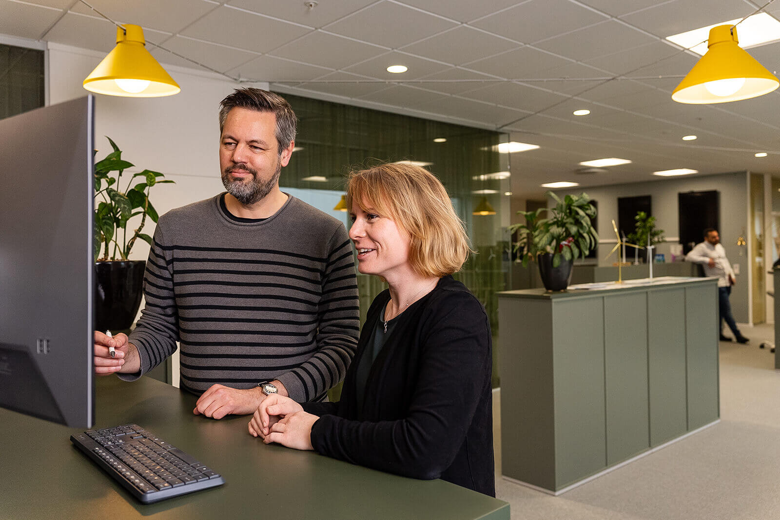 Two colleagues are discussing work at a desk with a computer and plants in a modern office environment.