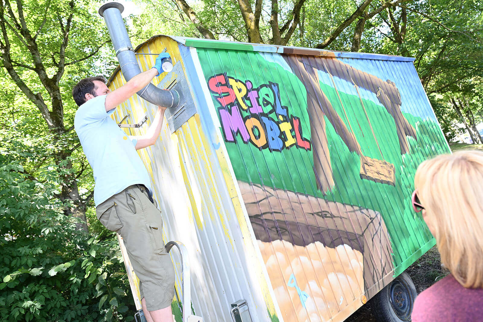 A man works on a brightly painted play vehicle, surrounded by trees and lush greenery.