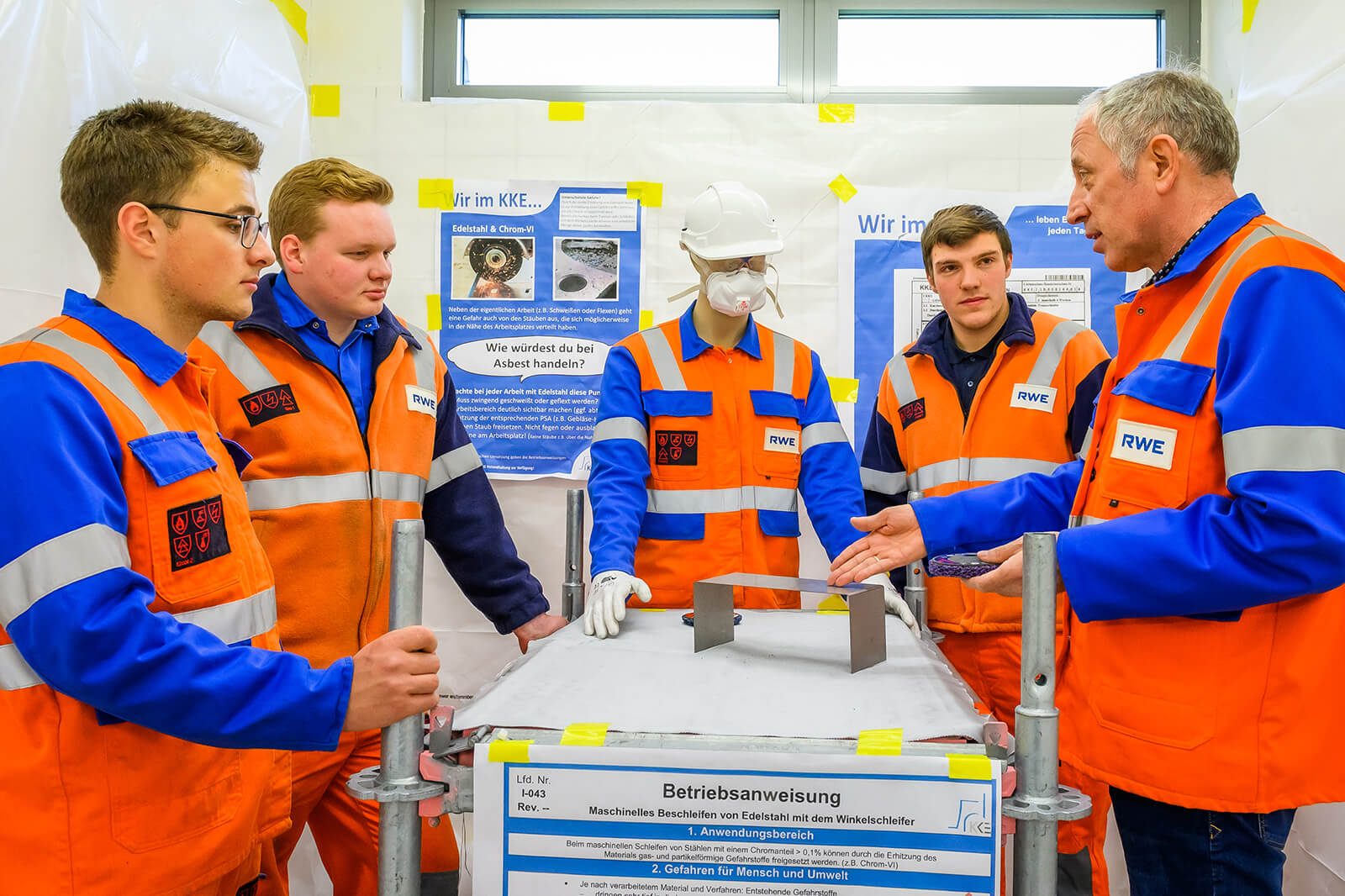 Five individuals in orange safety vests are discussing a metal object on a table.