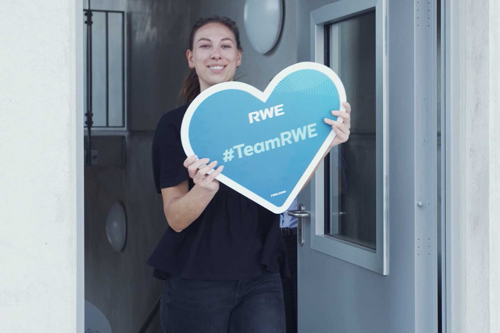 A person holding a heart-shaped sign saying RWE and #TeamRWE, standing at a doorway.